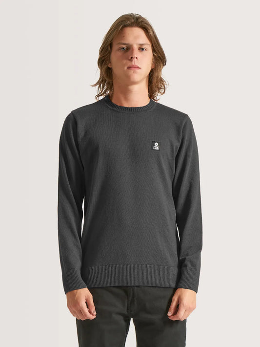 Homem vestindo blusa tricot preta, a peça possui barra, pulsos e gola em tecido canelado. Etiqueta preta e branca costurada no peito também compõe modelo.