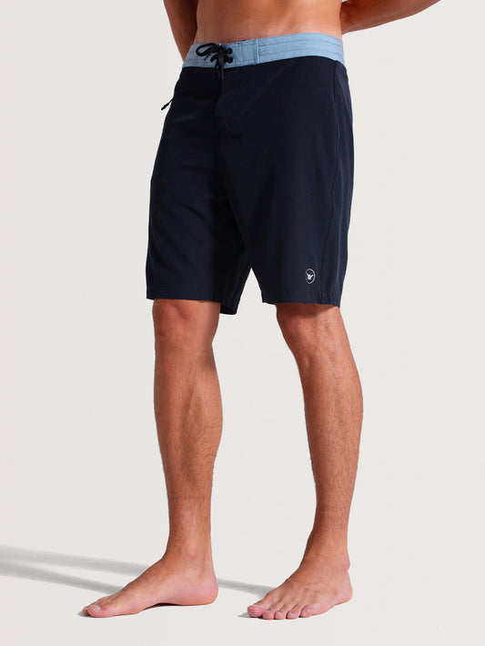 Homem vestindo boardshorts 19 polegadas azul com cós azul-claro contrastante, ajuste por cordão preto e design minimalista. Ideal para momentos casuais.
