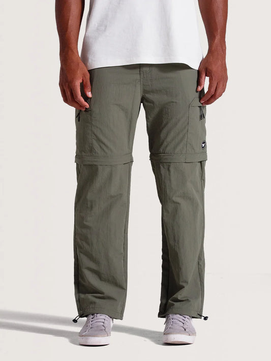 Homem vestindo calça híbrida 2 em 1 verde-oliva com ajuste por zíper para conversão em bermuda, bolsos laterais com zíper e cordões na barra. Estilo funcional.
