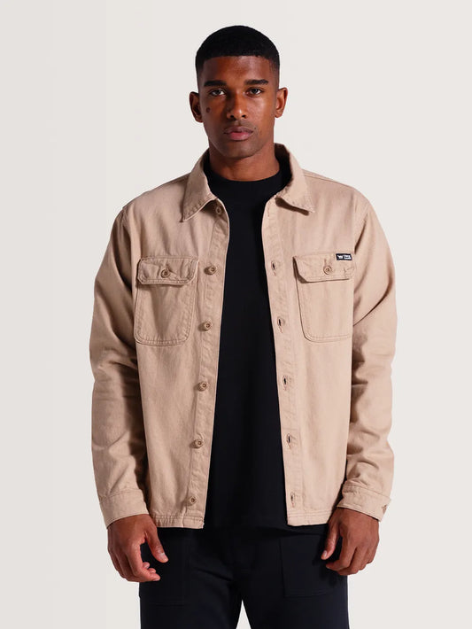 Modelo vestindo camisa sarja manga longa na cor bege. A peça possui bolsos frontais no peito e botões.