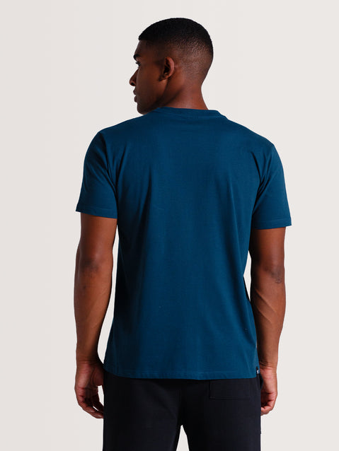 Homem de costas vestindo camiseta azul petróleo manga curta, combinada com calça preta, posando em fundo claro neutro.