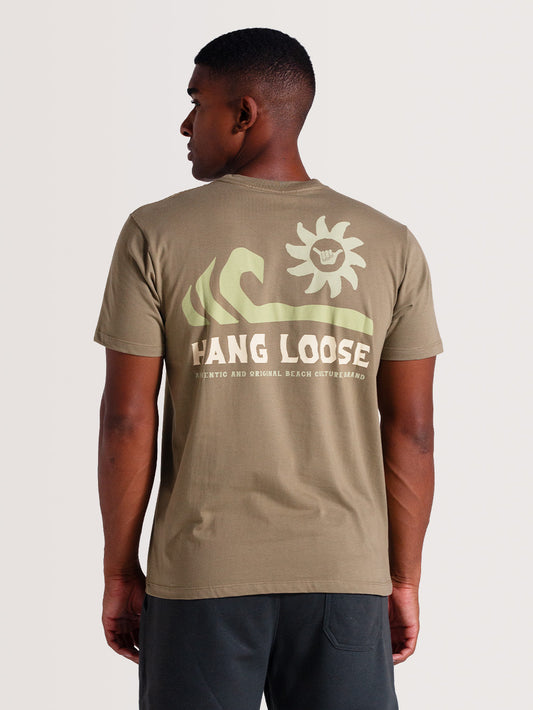 Homem de costas usando camiseta caqui manga curta estampada com design em tons claros de ondas, sol e texto "HANG LOOSE", posando contra fundo neutro.