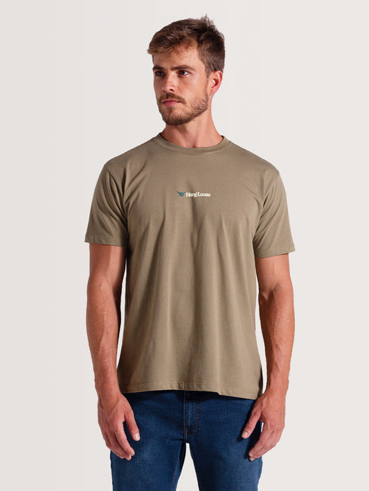 Homem vestindo camiseta caqui manga curta, com gola redonda e logo "Hang Loose" branco no peito, combinada com calça jeans azul, sobre fundo claro.