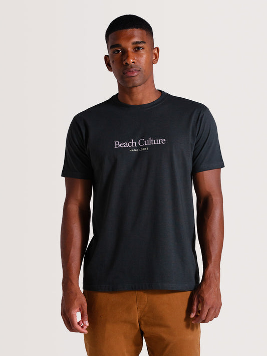 Homem vestindo camiseta cinza escuro manga curta com estampa "Beach Culture Hang Loose", combinada com calça bege, posando em fundo neutro.