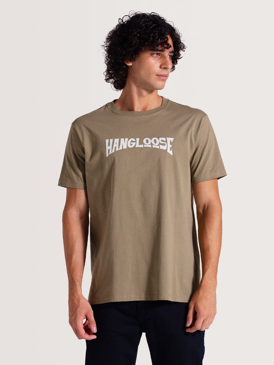 Homem vestindo camiseta cor caqui manga curta com estampa frontal branca "Hang Loose" em fonte estilizada, combinada com calça preta.