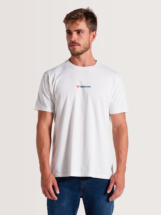Homem usando camiseta off white manga curta com gola redonda e logo "Hang Loose" no peito, combinada com calça jeans azul, fundo claro.