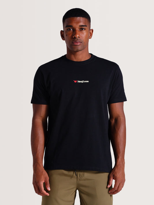 Homem vestindo uma camiseta preta manga curta com gola redonda e logo "Hang Loose" branco e vermelho no peito, combinada com calça caqui