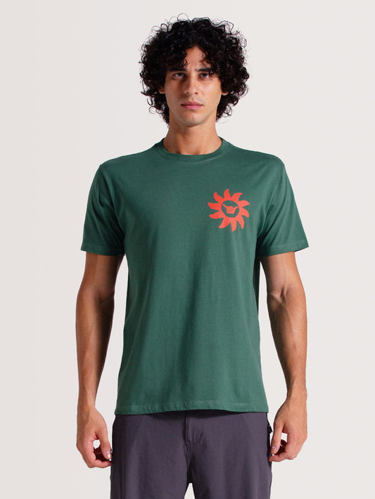 Homem vestindo camiseta verde musgo manga curta com estampa coral de um sol no lado esquerdo do peito, posando em fundo claro neutro.