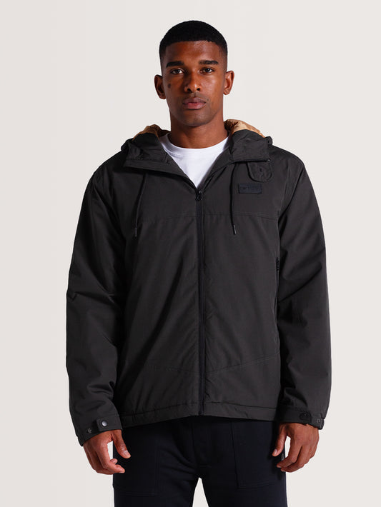 Homem vestindo jaqueta preta pesada com capuz de tecido robusto, com zíper frontal, detalhes minimalistas e forro interno bege visível no capuz.