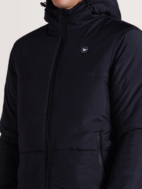 Close da jaqueta puffer preta acolchoada com capuz ajustável, zíper frontal e detalhe de pequeno emblema circular no peito.