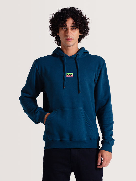 Homem vestindo moletom canguru azul com capuz, cordões ajustáveis e bolso frontal. Destaque para um pequeno patch colorido no peito. Fundo neutro.