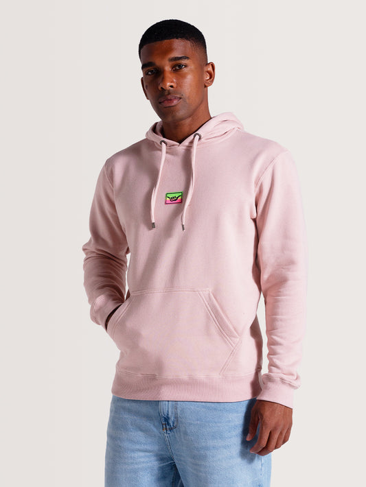 Homem vestindo um moletom canguru rosa com capuz, bolso frontal e cordões ajustáveis. Destaque para patch colorido no peito. Fundo claro e minimalista.