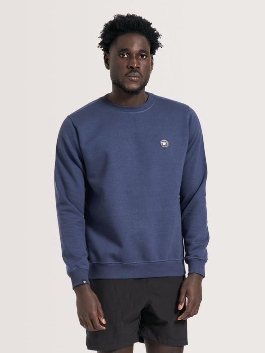 Homem vestindo moletom careca azul marinho com gola redonda, detalhe de mini logo circular da Hang Loose no peito esquerdo e tecido liso. Combinado com short preto casual.