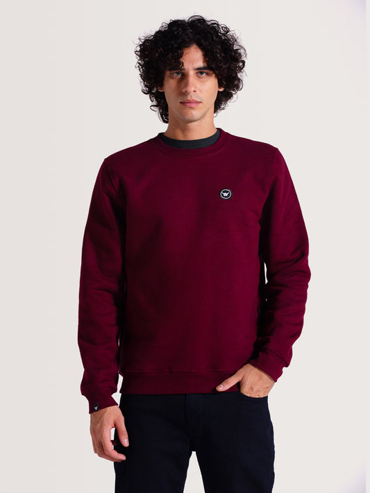 Homem veste moletom careca bordô com logo circular preto e branco no peito, ajuste confortável e discreto. Fundo claro destaca o tom intenso da peça.