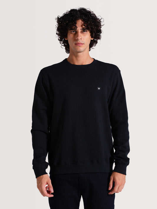 Homem vestindo o moletom careca preto básico, com mangas compridas e pequeno logotipo no peito. Fundo claro e visual casual e minimalista.