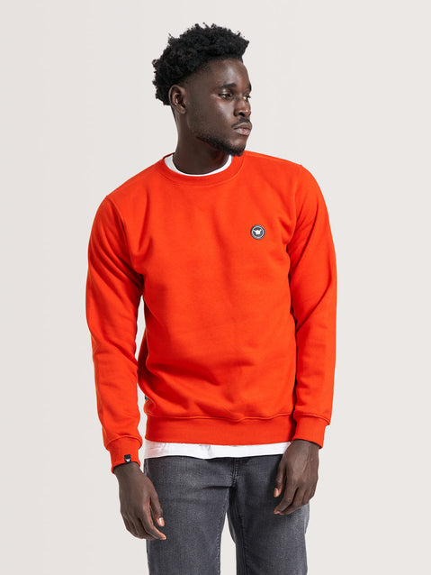 Homem vestindo moletom vermelho careca com gola redonda e ajuste regular. Moletom tem pequeno emblema no peito, combinado com jeans acinzentado.