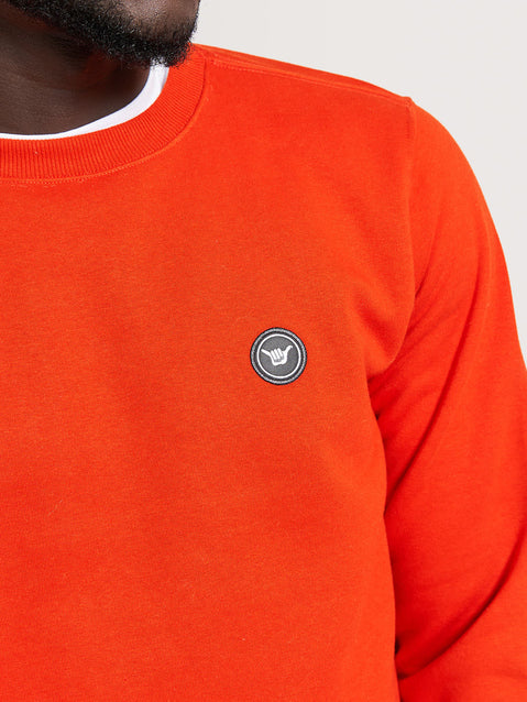 Homem usando moletom vermelho careca com gola redonda, modelagem simples e detalhe de emblema preto no lado esquerdo do peito.