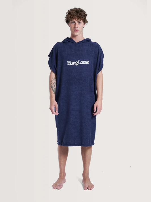 Modelo posando em fundo neutro vestindo o poncho surf da Hang Loose. A peça possui cor azul e nome da marca em branco na parte frontal.