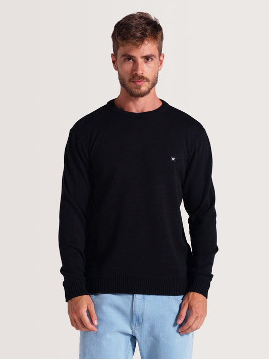 Homem vestindo tricot preto de manga longa, com gola redonda, combinado com calça jeans clara, fundo branco simples.