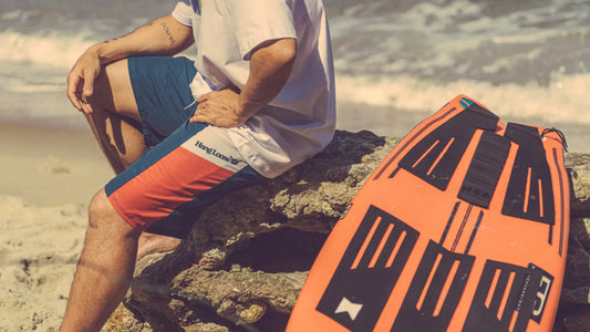 Imagem de homem vestindo boardshort e camiseta Hang Loose sentado em pedra na praia com prancha de surf ao lado.
