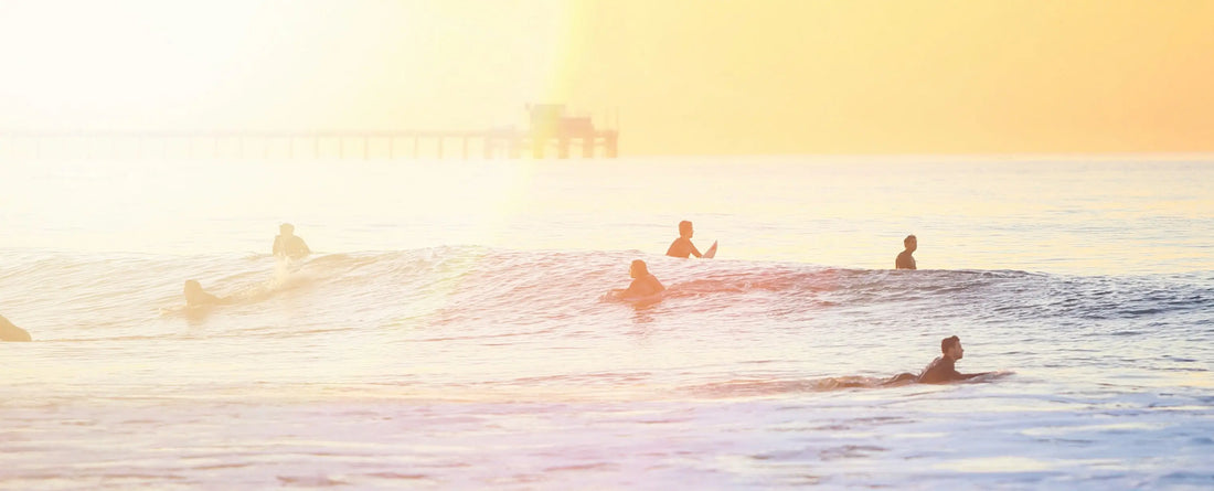 Surfistas no mar em cima de suas pranchas, ao fundo o céu está laranja e há um píer, o sol ilumina o lado esquerdo da imagem.