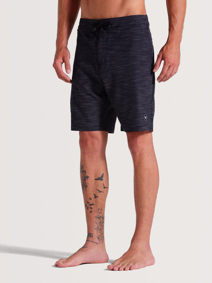 Homem vestindo bermuda híbrida 19 polegadas preto, estilo casual, com estampa discreta em tom mesclado e logo minimalista próximo à barra. Peça confortável e versátil.