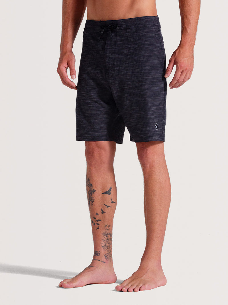 Homem vestindo bermuda híbrida 19 polegadas preto, estilo casual, com estampa discreta em tom mesclado e logo minimalista próximo à barra. Peça confortável e versátil.