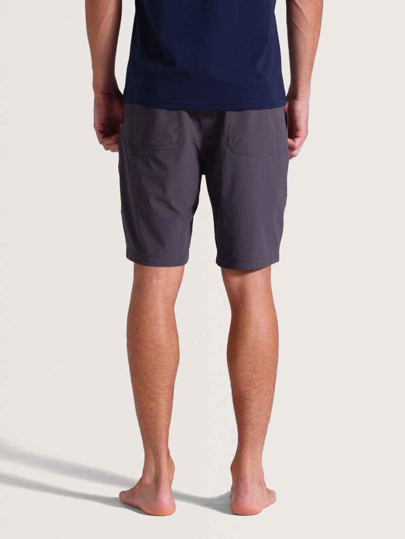 Homem de costas vestindo bermuda híbrida preta com bolsos traseiros simples, tecido leve e corte acima do joelho. Combina conforto e praticidade.