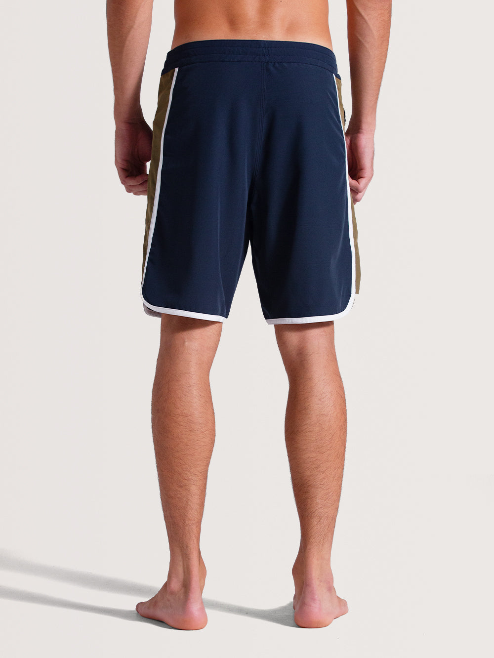 Homem de costas vestindo boardshorts 19 polegadas azul-marinho com detalhes laterais bege e brancos, barra arredondada e tecido leve, ideal para o verão.