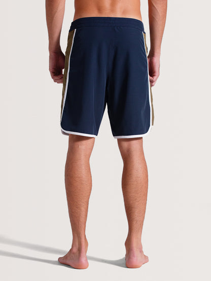 Homem de costas vestindo boardshorts 19 polegadas azul-marinho com detalhes laterais bege e brancos, barra arredondada e tecido leve, ideal para o verão.