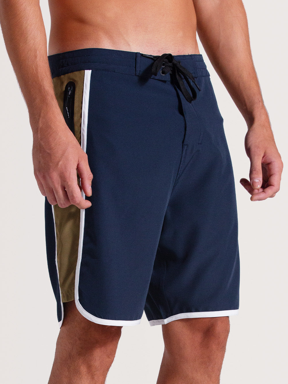 Homem vestindo boardshorts 19 polegadas azul-marinho com barra e detalhes brancos, bolso lateral bege com zíper e ajuste por cordão preto. Estilo esportivo e casual.