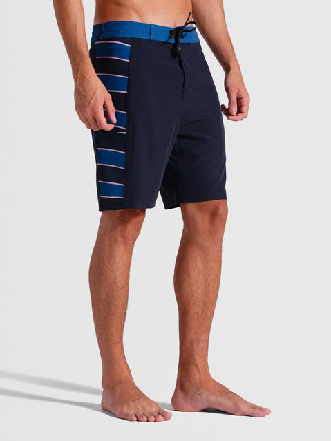 Homem vestindo boardshorts 19 polegadas chumbo com detalhes listrados em azul na lateral, cós azul contrastante e ajuste com cordão preto. Estilo esportivo e moderno.