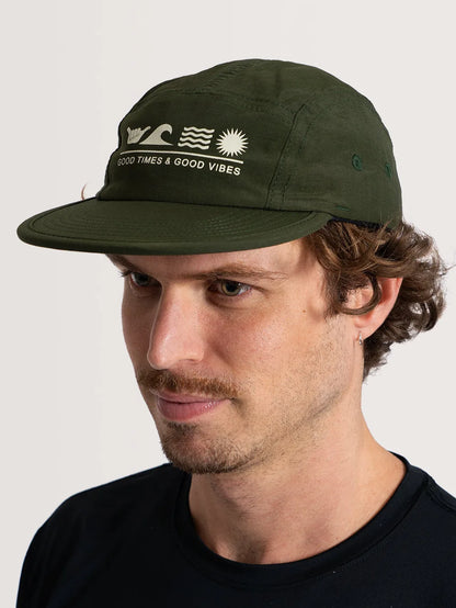 Homem usando boné verde com escrito "Good Times & Good Vibes" e símbolos de uma ondas, sol e o shaka, transmitindo uma vibe positiva.