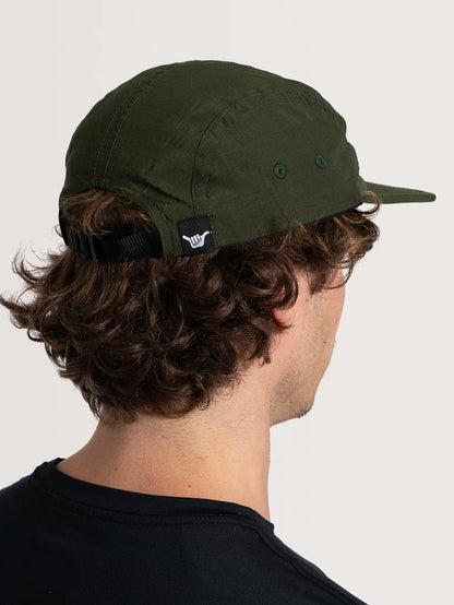 Homem usando o boné Hang Loose flat verde ao fundo branco, de perfil, com cabelo ondulado castanho, vestindo camiseta preta. Item no estilo five panel com etiqueta com símbolo shaka na lateral.