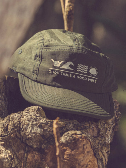 Chapéu verde militar Hang Loose flat com frase "Good Times & Good Vibes" em branco e símbolos de ondas, sol e shaka, colocado sobre um tronco de árvore.