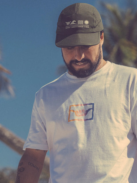 Homem com barba usando o boné flat Hang Loose verde militar e camiseta branca ao ar livre, com fundo de céu azul e árvores leves. O boné tem a frase "Good Times & Good Vibes".