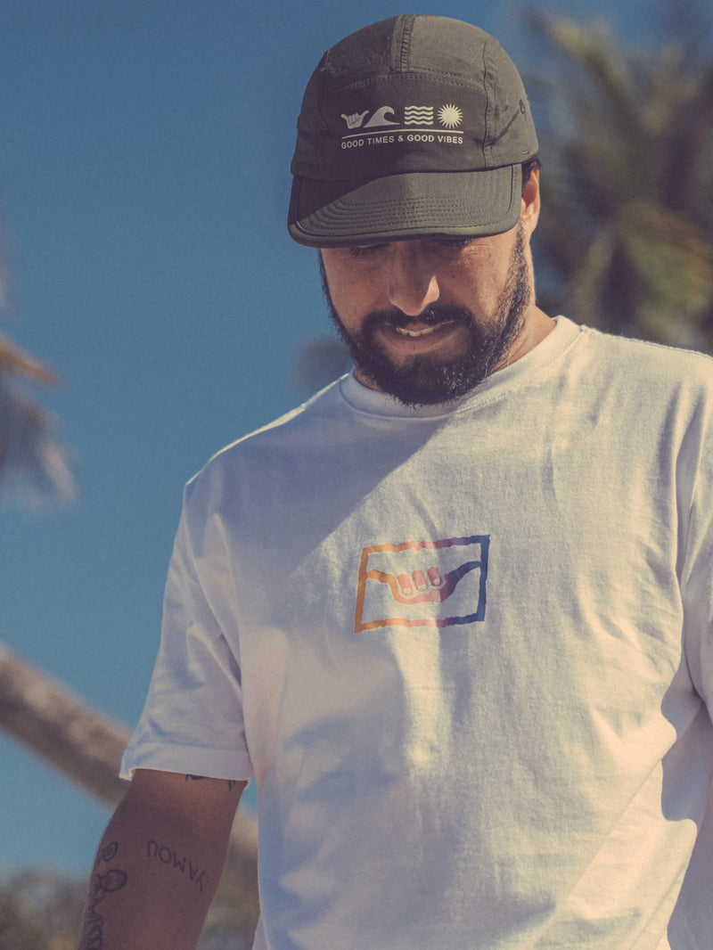 Homem com barba usando o boné flat Hang Loose verde militar e camiseta branca ao ar livre, com fundo de céu azul e árvores leves. O boné tem a frase "Good Times & Good Vibes".
