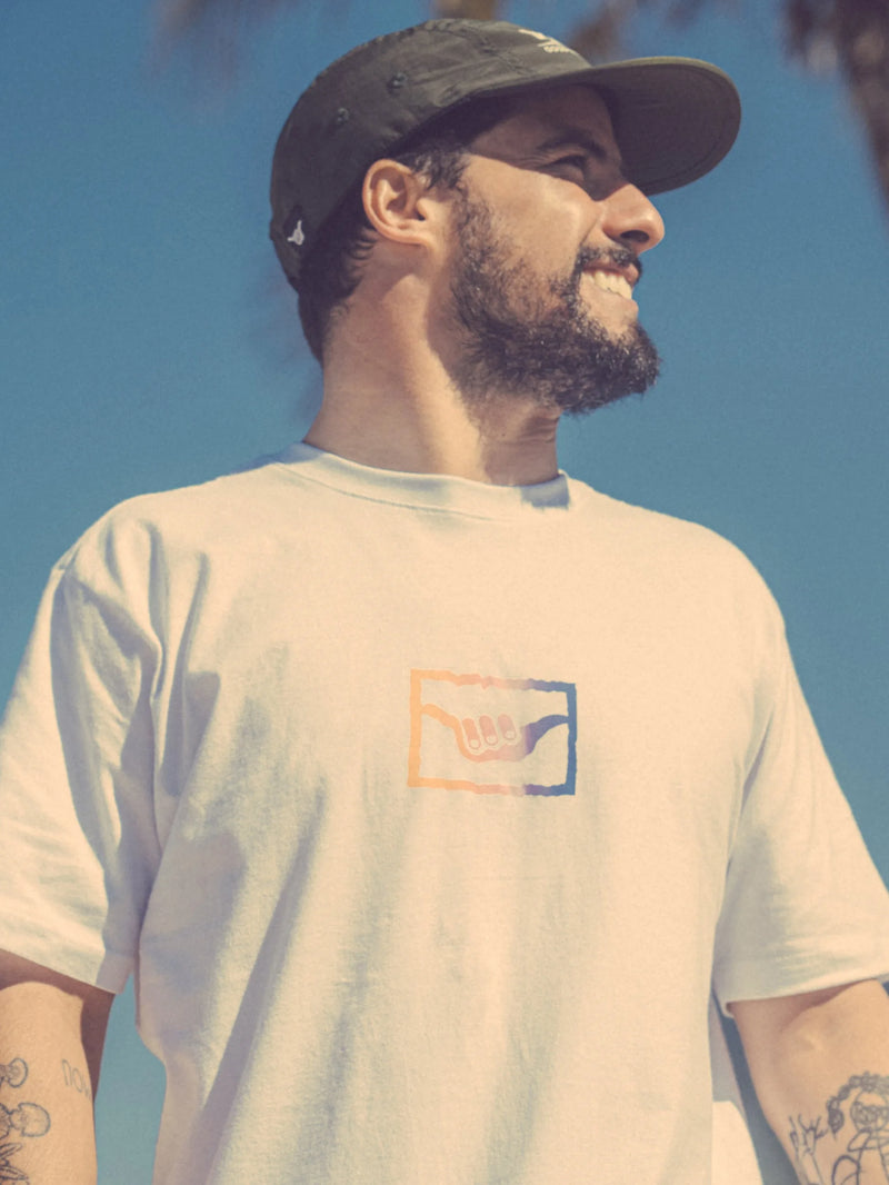 Modelo sorrindo ao ar livre usando boné Hang Loose Flat em estilo five panel e camiseta com símbolo shaka, com céu azul ao fundo, transmitindo sensação de liberdade e alegria.
