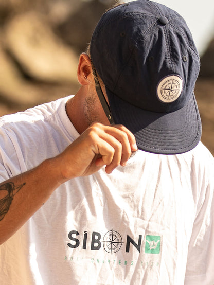 Close de homem ajustando boné azul marinho com logo de bússola, combinando com camiseta branca Sibon, ideal para estilo casual e aventuras ao ar livre.