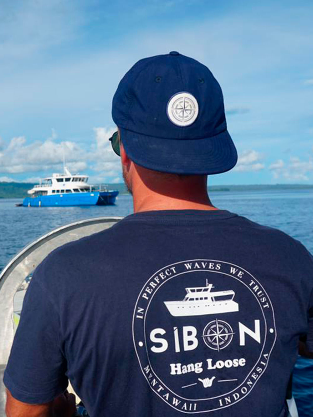 Homem usando boné azul marinho com logo de bússola, apreciando o mar ao lado de barco Sibon, ideal para aventuras e lifestyle náutico.