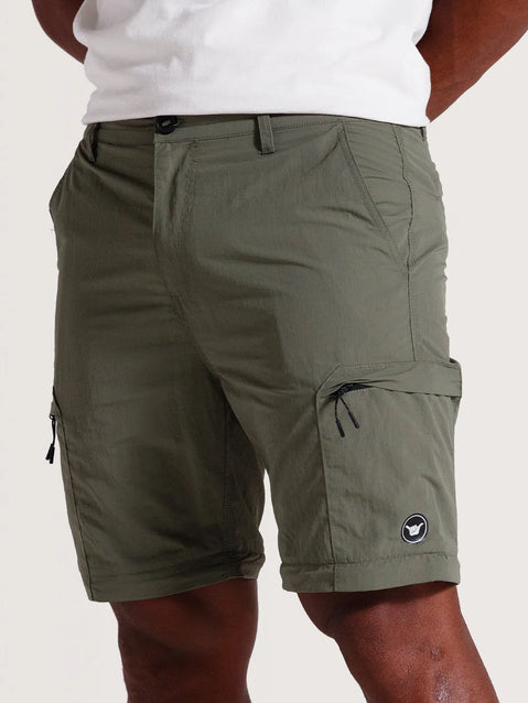 Modelo vestindo calça híbrida 2 em 1 convertida em bermuda verde, com bolsos laterais com zíper e pequeno logo em preto e branco próximo à barra.