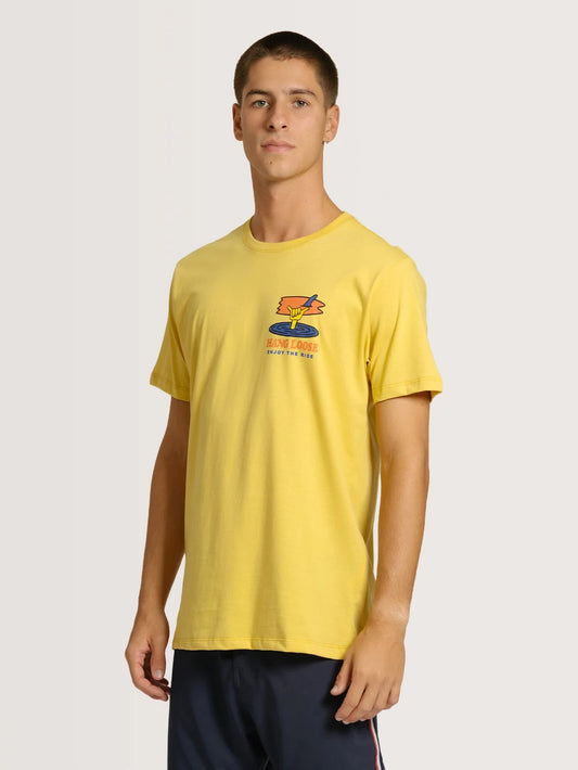 Homem vestindo camiseta amarela básica em fundo neutro. A peça possui arte nas cores amarelo, laranja e azul no peito esquerdo.
