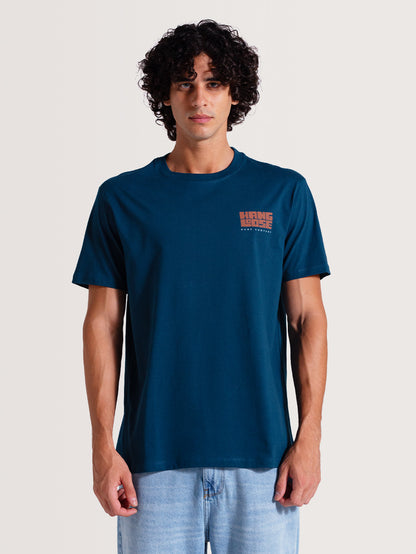 Homem vestindo camiseta azul manga curta com pequeno logotipo laranja no lado esquerdo do peito. Estilo casual e descontraído, combinado com jeans claro.