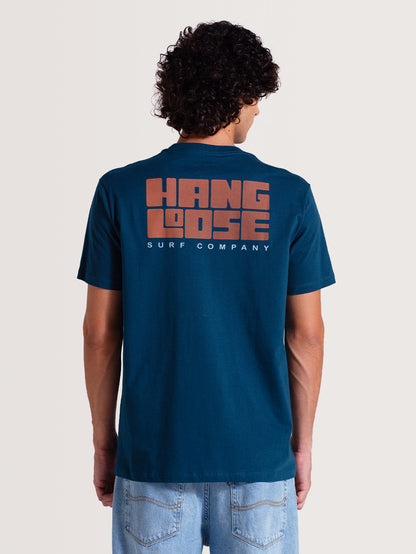 Homem vestindo camiseta azul manga curta com logo grande em laranja nas costas escrito "Hang Loose Surf Company". Combinada com jeans claro, estilo casual.
