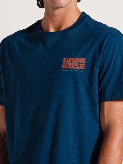 Homem vestindo camiseta azul manga curta com logo "Hang Loose Surf Company" pequeno em laranja no lado esquerdo do peito. Estilo casual e descontraído.