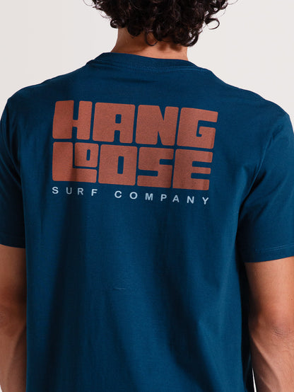 Camiseta azul manga curta com estampa grande "Hang Loose Surf Company" em laranja e branco nas costas. Estilo descontraído e casual.