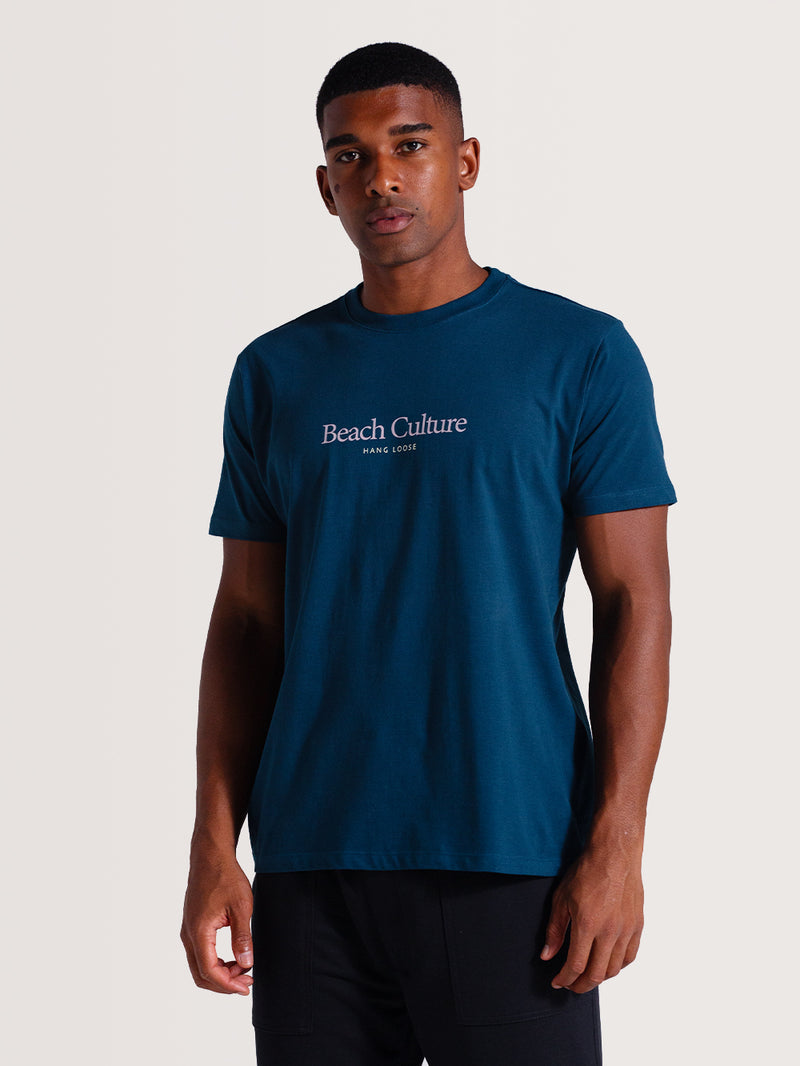 Homem usando camiseta azul petróleo manga curta com estampa central "Beach Culture Hang Loose", combinando com calça preta, posando em fundo neutro.
