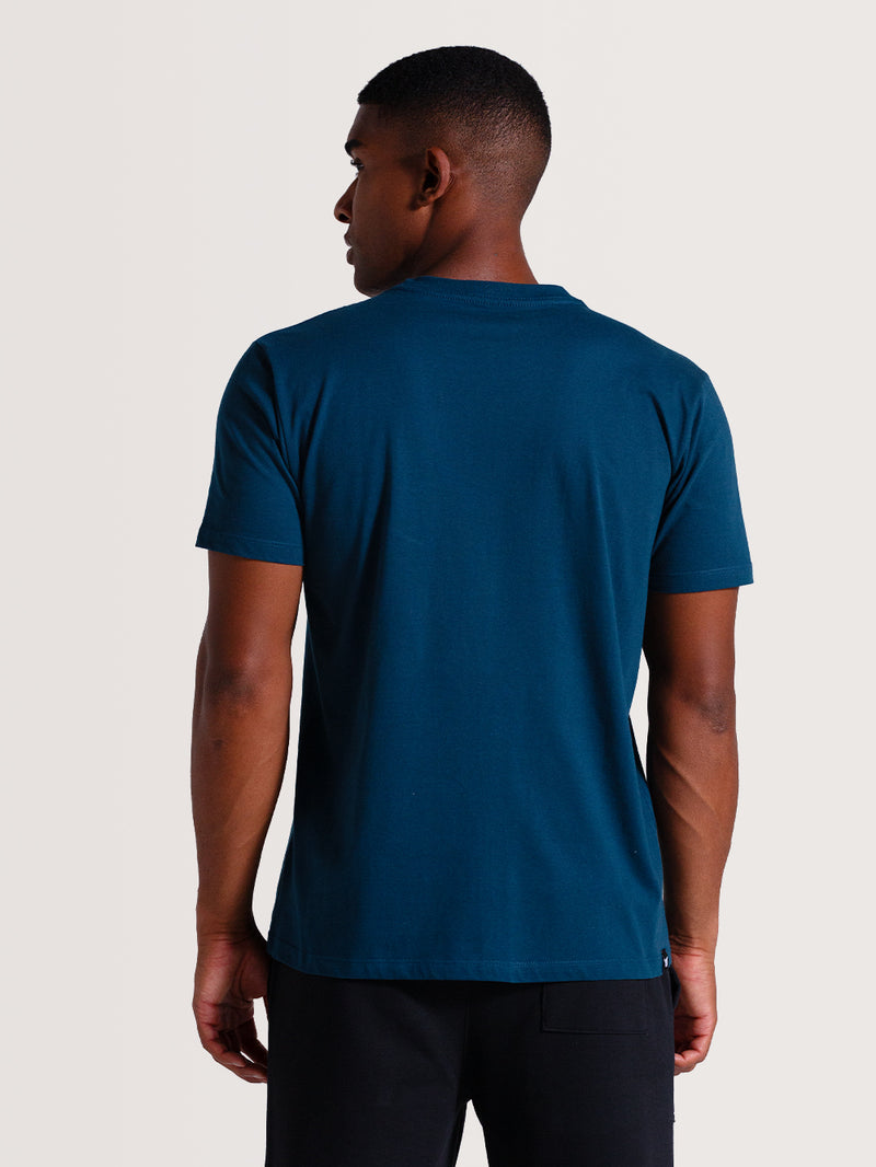 Homem de costas vestindo camiseta azul petróleo manga curta, combinada com calça preta, posando em fundo claro neutro.