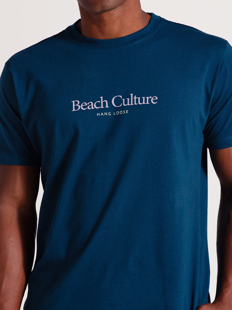 Homem vestindo camiseta azul petróleo manga curta com estampa central "Beach Culture Hang Loose", fotografado em plano médio contra fundo claro.