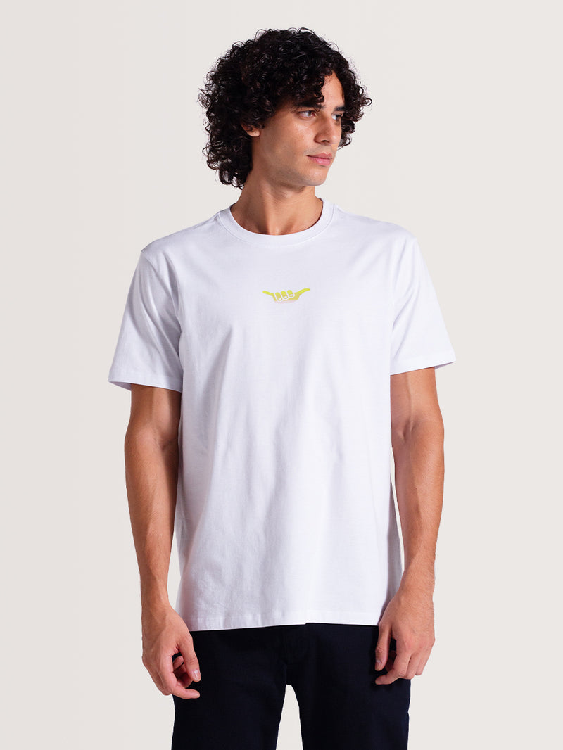 Homem vestindo camiseta básica branca manga curta com pequeno símbolo "Hang Loose" em amarelo no centro do peito, combinando com calça preta.